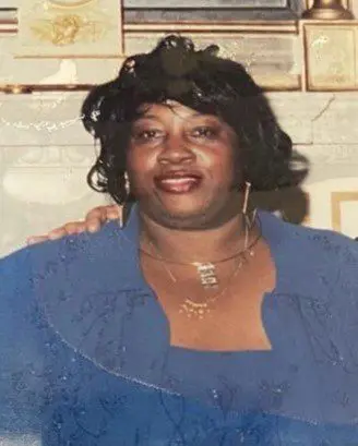 A woman in blue shirt and necklace standing next to wall.